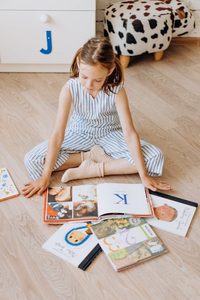 A cute child engaged in reading colorful books while sitting on a wooden floor indoors.